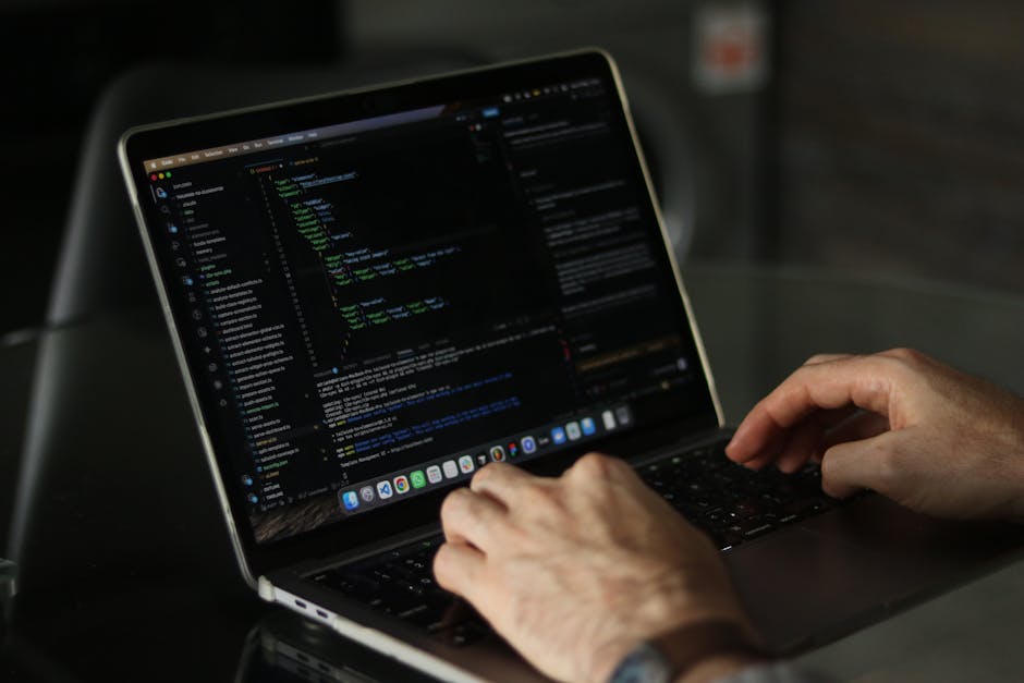 A person using a laptop on a dark table, focusing on a screen displaying a coding interface with lines of programming in various colors. The individual's hands are visible, with one hand poised over t
